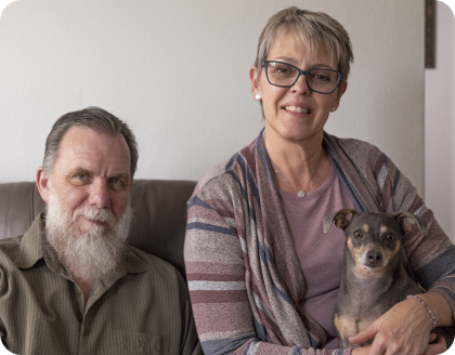 A smiling couple relaxing on a couch. The man has a full white beard, and the woman is holding a small, gray and tan dog in her lap.