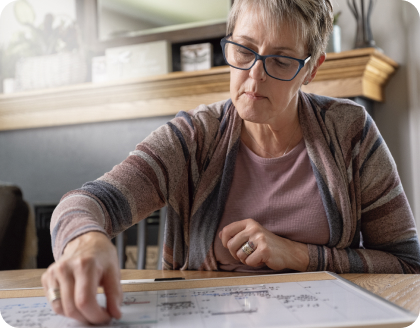 A middle-aged woman with short hair and glasses, wearing a striped cardigan, pointing to a document on a wooden table.